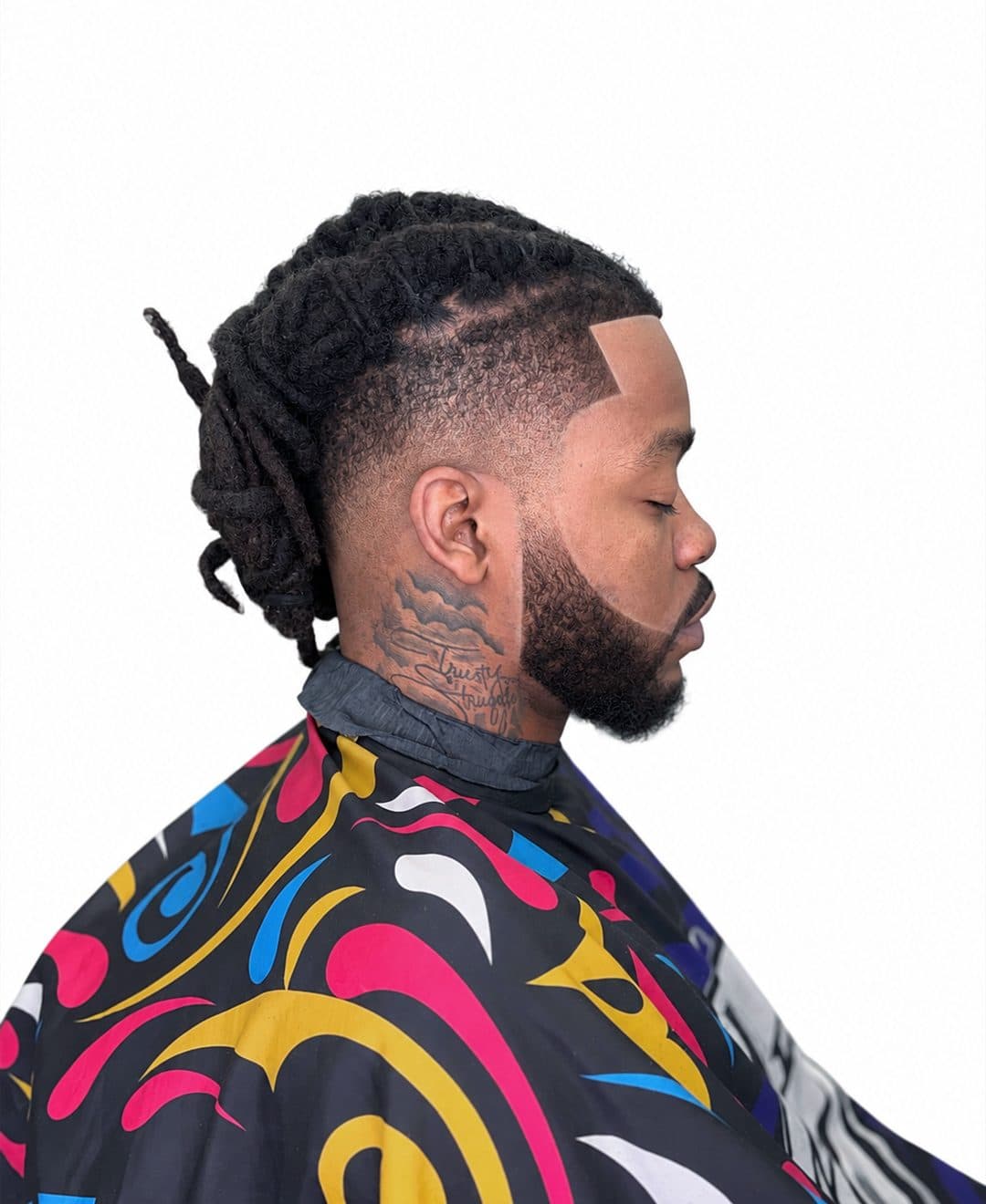 Locs recogidos sobre un high skin fade con línea marcada y barba completa, visto de perfil